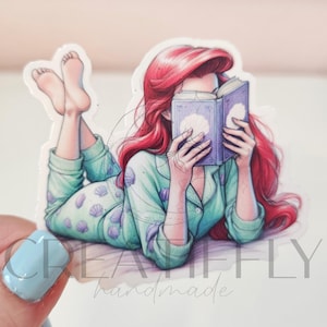 May include: A sticker featuring a cartoon illustration of a person with red hair reading a book while wearing light green pajamas with seashell patterns. The person is lying down with their feet up.