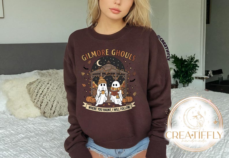 May include: A dark brown sweatshirt with the text "GILMORE GHOULS" above an illustration of two ghosts in a gazebo. The ghosts are holding a cup and a book. The text "WHERE YOU HAUNT, I WILL FOLLOW" is below the illustration.