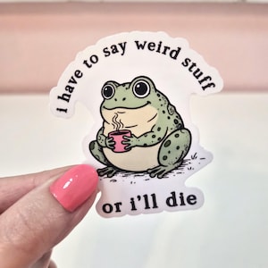 May include: A die-cut vinyl sticker featuring a light green frog with dark spots holding a small pink mug with steam rising from it. The text "i have to say weird stuff or i'll die" is printed in black, curved around the top and bottom of the frog.