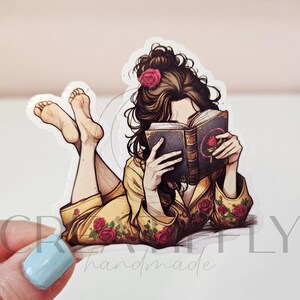 May include: A sticker featuring a person lying down and reading a book. The person is wearing a yellow robe with red rose accents and a pink rose in their hair. The book has a rose design on the cover.