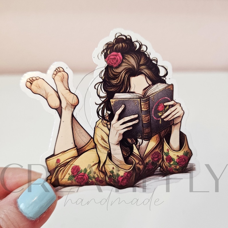 May include: A sticker featuring a person lying down and reading a book. The person is wearing a yellow robe with red rose accents and a pink rose in their hair. The book has a rose design on the cover.