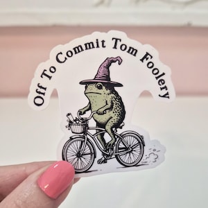 May include: A die-cut sticker featuring a green frog wearing a purple pointed hat, riding a bicycle with a front basket containing a scroll. The text "Off To Commit Tom Foolery" is arched above the frog in a black, serif font on a white background.