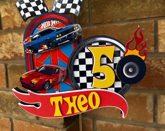 May include: A colorful birthday cake topper featuring a racing theme. It includes a blue car with flames, a red car, checkered flags, and the number 5. The name "Theo" is also displayed in yellow letters on a red banner.