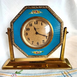 May include: A gold-toned clock with a blue face and floral accents. The clock is on a gold stand and has Roman numeral markings.