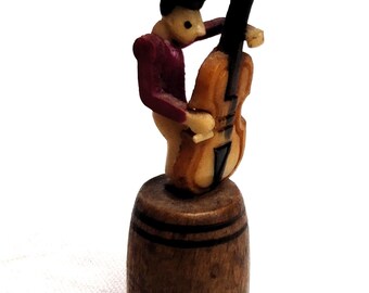 May include: A wooden figurine of a person playing a cello, standing on a wooden barrel. The figurine is painted in shades of brown, red, and green.