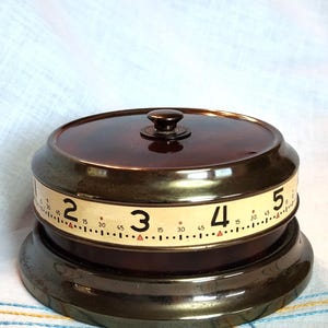 May include: A vintage brown and brass kitchen timer with a rotating dial. The timer features a dark brown top with a small knob and a cream-colored band with black numbers indicating minutes. The base is a darker brass color.