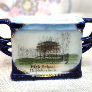 May include: A small, square, blue and white ceramic cup with two handles. The cup features a painted scene of a building and trees, with the text "High School Mapleton Iowa". Gold trim accents the edges.