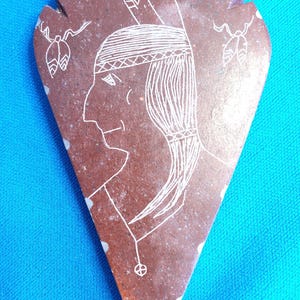 May include: A brown, arrowhead-shaped stone carving featuring a Native American profile. The white etched design includes a feathered headdress, long hair, and decorative elements. The background is a solid blue.