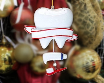 May include: A white tooth-shaped ornament with a glittery finish, featuring a blank white banner with red trim wrapped around the center. Below the tooth, a small white toothpaste tube and a red toothbrush are suspended by a silver link.