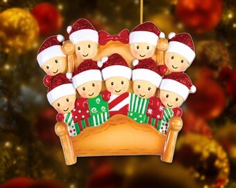 May include: A Christmas tree ornament featuring eight cartoon figures with smiling faces, each wearing a red glittery Santa hat. The figures are arranged in two rows behind a light brown wooden headboard with a red bow at the top center.