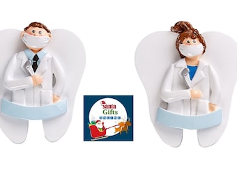 May include: Two white tooth-shaped Christmas decorations with angel wings. Each decoration features a dentist wearing a white coat, a face mask, and holding a dental tool. The decorations are decorated with a blue ribbon.