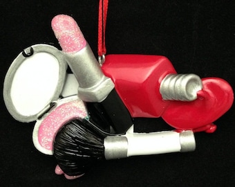 Könnte beinhalten: Ein rosa und silberner Lippenstift, ein roter Nagellackflakon, eine rosa und silberne Kompaktdose mit Spiegel und ein schwarzer Make-up-Pinsel mit silbernem Griff. Die Artikel sind in einer Gruppe angeordnet und an einem roten Band befestigt.