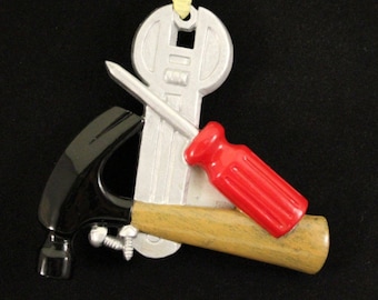 Könnte beinhalten: Ein silberner Schraubenschlüssel, ein roter Schraubendreher und ein schwarzer Hammer mit einem Holzgriff sind in einem dekorativen Design angeordnet. Die Werkzeuge sind an einem gelben Band befestigt, was auf einen Weihnachtsschmuck hindeutet.