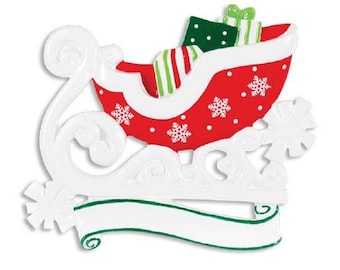 Puede incluir: Adorno navideño blanco y rojo con forma de trineo con regalos en su interior. El trineo tiene un patrón de copos de nieve blancos y puntos rojos. El adorno tiene una pancarta en blanco para personalizar.