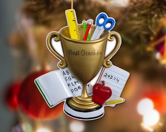 May include: A gold-colored trophy ornament featuring the text Best Teacher, containing a yellow ruler, a green pencil, a red pencil, and blue scissors. An open book with letters and a paper with 2+2=4 and a red apple are attached.