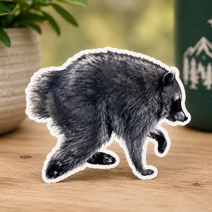 May include: A die-cut sticker featuring a detailed illustration of a raccoon in a walking pose. The raccoon is rendered in shades of black, gray, and white, showcasing textured fur, a pointed snout, and a characteristic dark mask around its eyes.