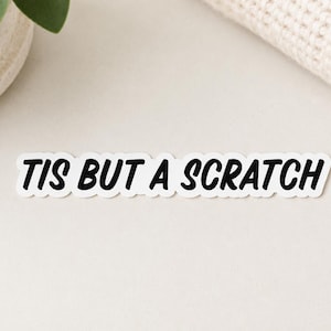 May include: A rectangular die-cut sticker featuring the black text TIS BUT A SCRATCH in a bold, sans-serif font. The sticker has a white background and a thin white border that follows the contour of the lettering, creating a clean, high-contrast look.