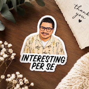 May include: A die-cut sticker featuring a cartoon illustration of a person with short dark hair, a mustache, and dark-rimmed glasses, wearing a beige short-sleeved shirt with a brown floral pattern, with the text INTERESTING PER SE below.