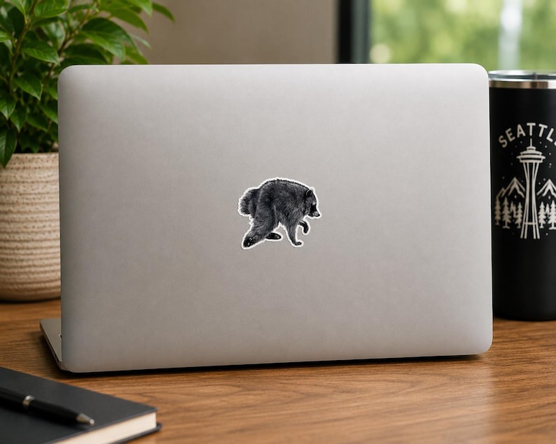 May include: A silver laptop featuring a centered sticker of a black and white raccoon illustration. The raccoon is depicted in a hunched, walking posture with detailed fur texture. The laptop lid is smooth and metallic, providing a neutral backdrop.