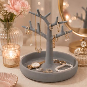 May include: A matte gray jewelry stand shaped like a tree with multiple branches for hanging necklaces, earrings, and rings. The base is a circular tray holding a gold-tone wristwatch, a pearl bracelet, and several gold rings and chains.