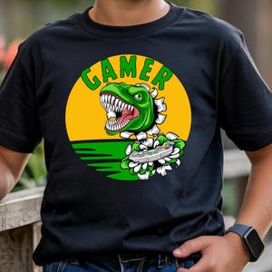 May include: A black short-sleeve t-shirt featuring a graphic of a green dinosaur breaking through a yellow circle. The dinosaur has sharp teeth and is holding a gray video game controller. The word GAMER is printed in green above the dinosaur.