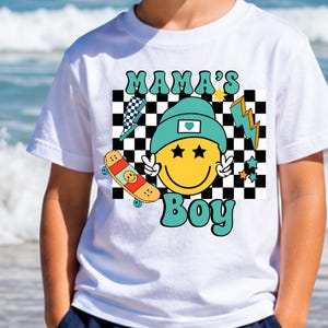May include: A white t-shirt featuring a graphic print with the text MAMA'S BOY in teal. The design includes a yellow smiley face wearing a teal beanie, a black and white checkered background, a skateboard, lightning bolts, and stars.