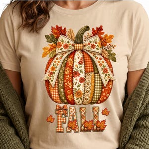 May include: A beige t-shirt featuring a graphic of a pumpkin made of various patterned fabric segments, topped with a large bow and autumn leaves. Below the pumpkin, the word FALL is written in block letters with a plaid pattern and leaf accents.