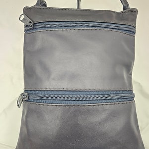 Puede incluir: Un bolso bandolera de cuero azul oscuro con dos compartimentos con cremallera y un asa pequeña. El bolso tiene forma rectangular y una textura suave. Las cremalleras son de color azul oscuro a juego.