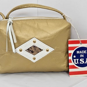 Puede incluir: Bolso rectangular dorado con asa y cremallera blanca. El bolso presenta un diseño en forma de diamante blanco y marrón con tachuelas doradas. Una etiqueta con el texto "MADE IN U.S.A." está adjunta.