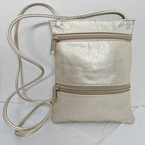 Puede incluir: Un bolso bandolera dorado metálico con dos compartimentos con cremallera y una correa larga y fina. El bolso es rectangular y tiene una superficie texturizada. Las cremalleras y la correa son doradas.