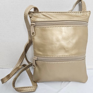 Puede incluir: Un bolso bandolera dorado con dos compartimentos con cremallera y una correa larga y fina. El bolso está hecho de un material suave y brillante y tiene un diseño simple y minimalista. Las cremalleras y la correa son del mismo color que el bolso.