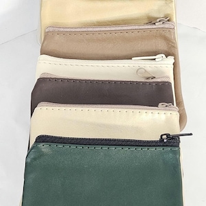 May include: A stack of small, zippered coin purses in various colors. The purses include shades of cream, gold, brown, and green. Each purse has a matching zipper and a stitched edge.