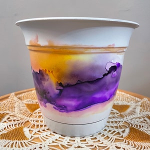 以下が含まれることがあります： 白い植木鉢にペイントされたデザイン。デザインは、黄色、紫、白などの色のブレンドが特徴で、抽象的で水彩画のような効果を生み出しています。鉢は、クリーム色の装飾的なドイリーの上に置かれています。