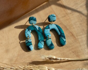 May include: A pair of abstract, arch-shaped earrings in shades of teal and blue, with a marbled effect. The earrings are suspended from small gold-colored posts and connected with gold-colored jump rings. They rest on a light wooden surface.