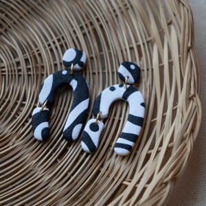 May include: A pair of black and white patterned earrings shaped like an arch. The earrings are made of polymer clay and have a gold-tone hook.