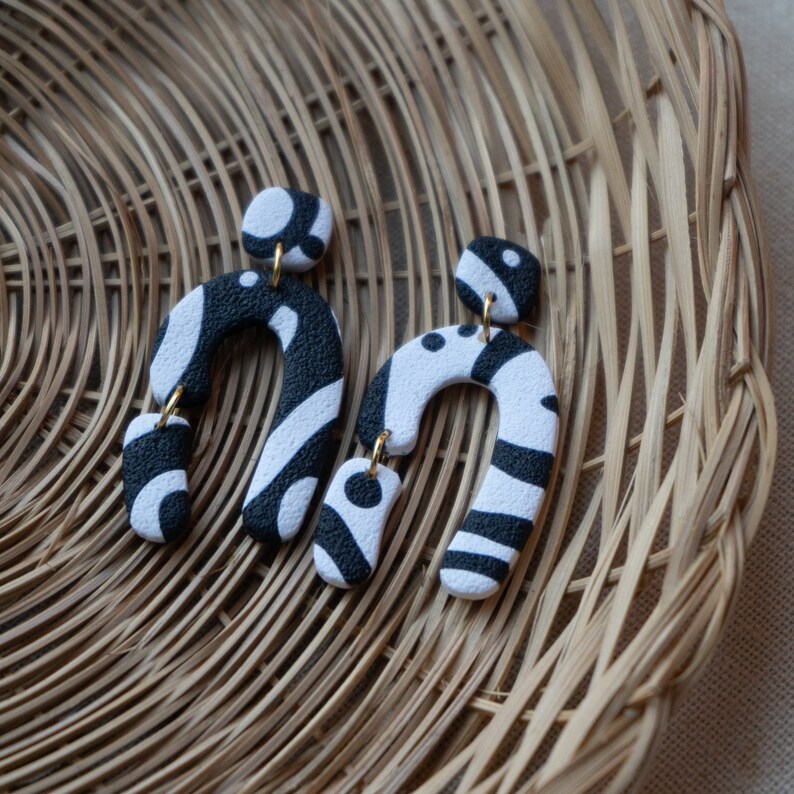 May include: A pair of black and white patterned earrings shaped like an arch. The earrings are made of polymer clay and have a gold-tone hook.