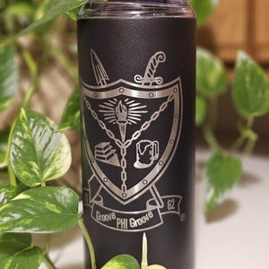 May include: Black stainless steel tumbler with a silver engraved design of a shield with a torch, a book, a mug, and swords. The text "Groove PHI Groove 62" is engraved on a banner below the shield.