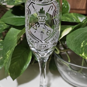 May include: Clear champagne flute with etched Greek letters and a crest design. The glass features a long stem and a wide base. The design includes a figure in profile, a torch, and the Greek letters Delta, Sigma, and Theta.