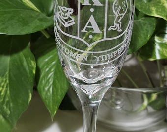 May include: A clear champagne flute with a decorative etched design. The design includes the letters A, K, and A within a shield, along with other symbols. The glass is set against a backdrop of green foliage.