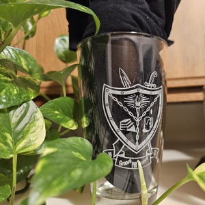 May include: A clear glass with a black graphic design featuring a shield, sword, and torch. The text "Groove Philosophy" is printed at the bottom. The glass is partially obscured by a black cloth and surrounded by green leafy plants.