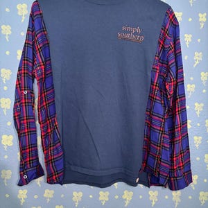 May include: A navy blue long-sleeved shirt with plaid sleeves in red, blue, and black. The shirt has the text "simply southern" in pink on the front. The shirt is hanging on a white hanger.