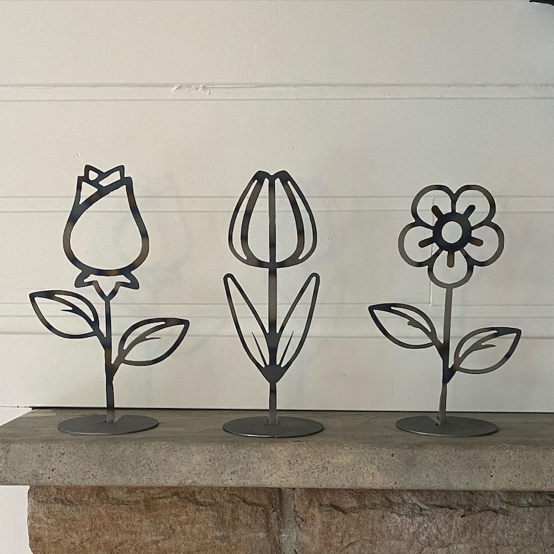 May include: Three metal flower sculptures on stands. The flowers are a rose, a tulip, and a daisy. The sculptures are black and are on a grey stone surface.