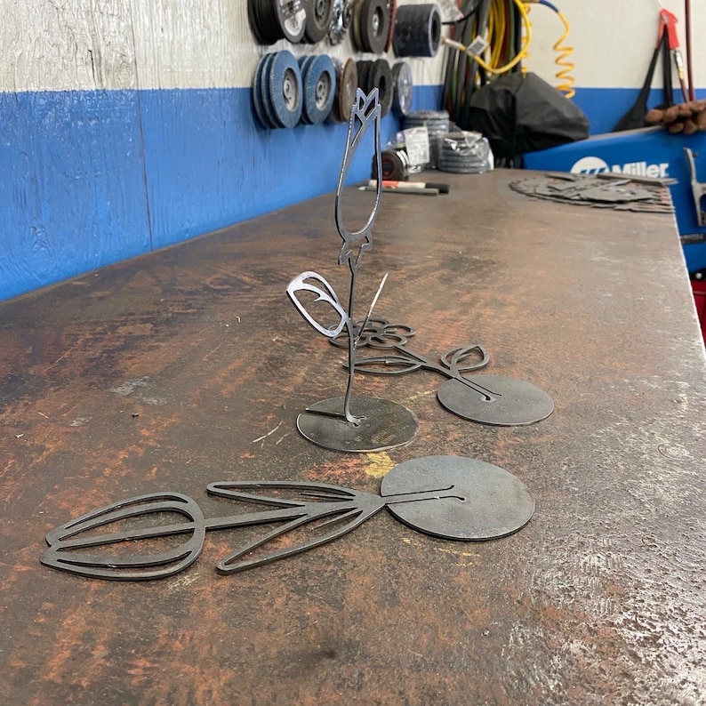 May include: Three metal flower sculptures with a black finish. The sculptures are of different sizes and feature a tulip, a rose, and a leaf design. The sculptures are sitting on a metal surface.