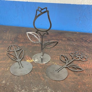May include: Three metal flower sculptures with black silhouettes on round metal bases. The flowers are a rose, a tulip, and a daisy.