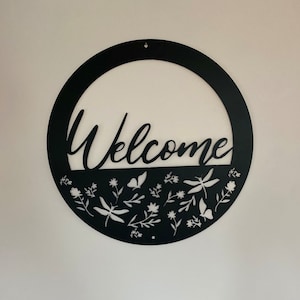 Op de afbeelding: Zwart metalen welkom bord met een cirkelvormig ontwerp. Het woord "Welkom" is uit het metaal gesneden en de onderste helft van de cirkel heeft een bloemmotief met witte uitsnijdingen van bloemen, bladeren en libellen.