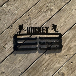 Op de afbeelding: Zwarte metalen hockeymedaillehouder met twee hockeyers en het woord "HOCKEY" in het midden. De houder heeft drie rijen haken voor medailles.