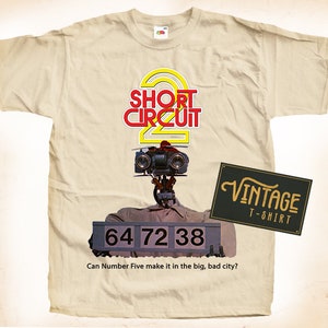 Può includere: T-shirt color crema con grafica del film "Corto Circuito 2". Il design include il titolo del film, un'immagine di robot e il testo "64 72 38". È presente anche un'etichetta vintage per t-shirt.