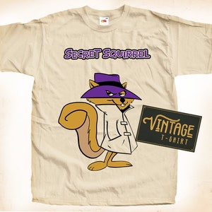 May include: A cream-coloured t-shirt featuring the cartoon character Secret Squirrel. The design includes the text "SECRET SQUIRREL" in purple and a vintage-style graphic. The squirrel is wearing a purple hat and trench coat.