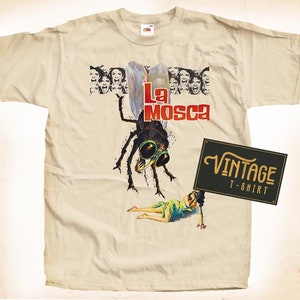 May include: Cream-coloured vintage t-shirt with a movie poster design. The graphic shows a large fly-like creature, the text "La Mosca" in red, and a woman in a green dress. A black sign reads "Vintage T-Shirt."