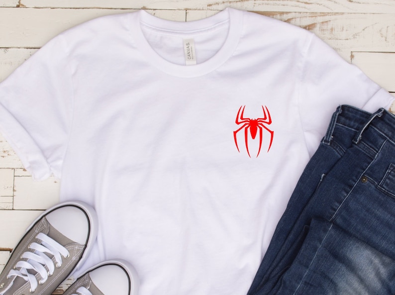 May include: White t-shirt with a red spider-man logo on the front. The shirt is laying on a white wooden surface with a pair of gray sneakers and blue jeans.